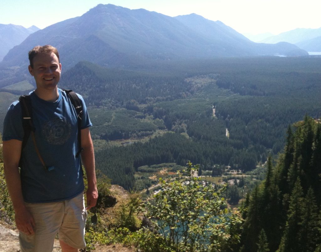 James Amadon stands on Rattlesnake Ledge with a small mountain range in the distance and a forested valley floor just behind him.