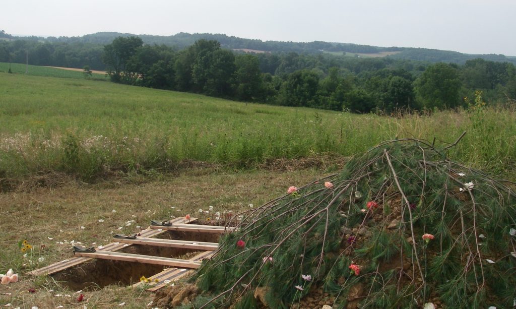 In rolling Ohio landscape hole for burial has been opened in meadow with pine branches nearby for covering after burial.