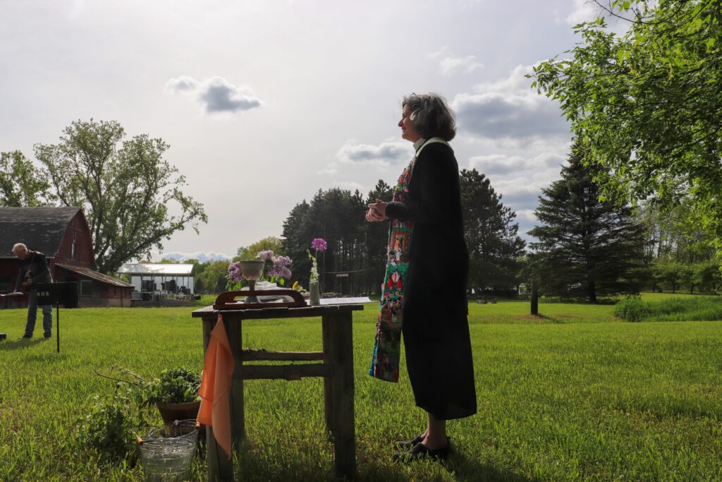Reverend Nurya Love Parish stands at outdoors altar at Plainsong Farm with trees and bright sky in background